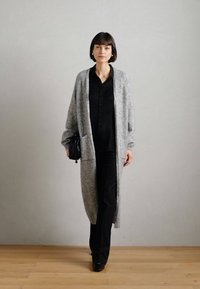 Cárdigan largo de punto gris con apertura frontal, que presenta mangas largas y un corte relajado. Lleva puesto sobre una camisa negra de botones y pantalones negros.