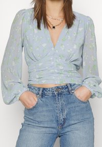 Kvinna som bär en ljusblå blommig V-hals blus med puffärm och högmidjade ljusblå jeans med händerna i fickorna.