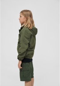 Veste à capuche verte avec des poignets élastiques, associée à un short cargo vert. Texture lisse et coupe décontractée, présentée de côté.