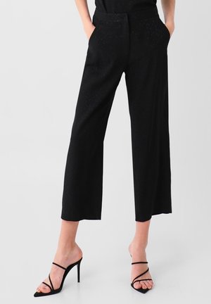 Pantalon noir large raccourci, taille haute avec poches, porté avec des sandales noires à talons hauts et à brides, mains dans les poches.