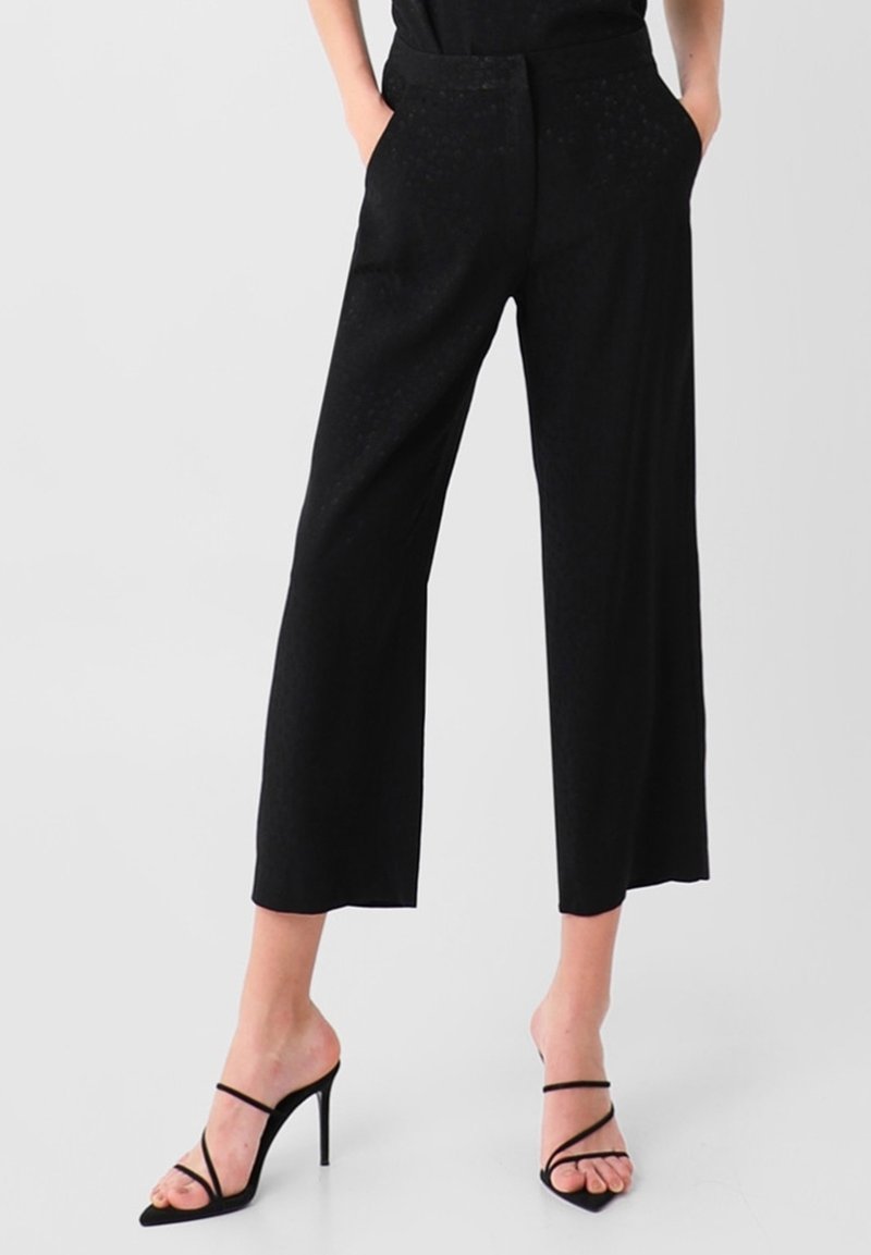 Pantalon noir large raccourci, taille haute avec poches, porté avec des sandales noires à talons hauts et à brides, mains dans les poches.