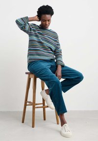 Pullover a strisce multicolore con pantaloni in corduroy blu e sneakers bianche. Modello seduto su uno sgabello di legno, con materiali testurizzati visibili.