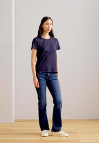 Ciemnoniebieska koszulka z krótkim rękawem w połączeniu z dopasowanymi ciemnymi dżinsami i białymi trampkami. Koszulka ma prosty, klasyczny fason z okrągłym dekoltem.