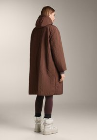 Smeđi kaput sa kapuljačom, stezane pamučne tkanine, s udobnim krojem i vidljivim šavovima. Uparen s ljubičastim tajicama i bijelim čizmama sa krznom.