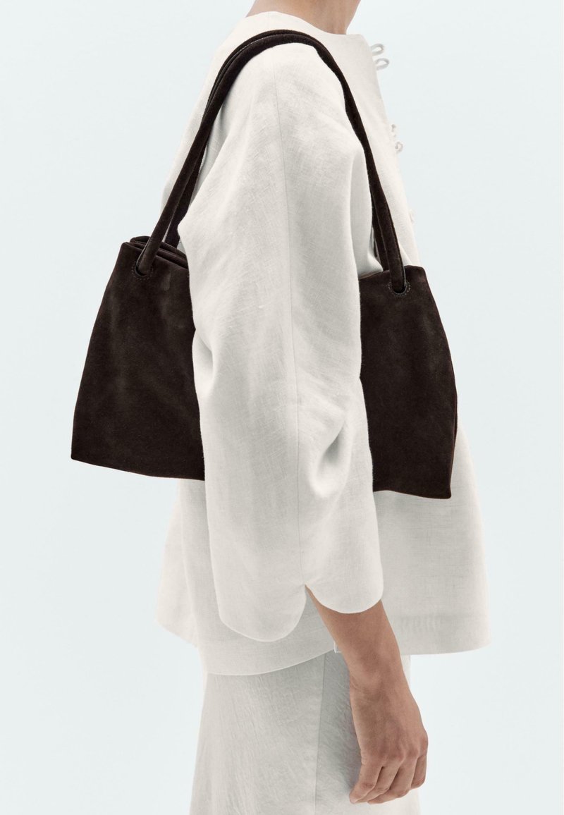 Sac tote en suède marron foncé avec des bords arrondis, équipé de deux longues poignées et d'un design minimaliste. Porté par un mannequin dans une tenue blanche.