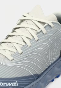 Baskets de sport gris clair avec texture nervurée, lacets blancs et semelle bleue. Présente des motifs ondulés sur le côté pour un détail de design supplémentaire.
