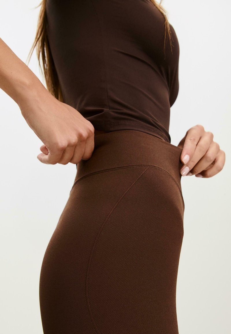 Osoba prilagođava pojas smešnih pantalona, noseći odgovarajući smešni top, na jednostavnoj svetloj pozadini.