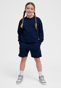 Navyblauwe sweatshirt en short set in zachte stof. Sweatshirt heeft een klein logo-embleem; shorts beschikken over zijzakken. G gedragen met grijze sneakers.