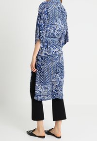 Kobieta w niebieskiej, wzorzystej długiej kurtce kimono, nosząca czarne spodnie, 3/4 i czarne, ćwiekowane buty wsuwane, stojąca na tle w neutralnym kolorze.