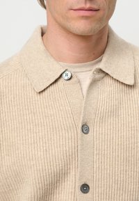 Beige ribbed knit shirt jacket with a collared neckline, two gray buttons, and a soft texture. Ideal for layering in casual settings.