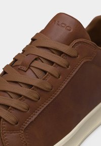 Baskets en cuir marron avec lacets assortis, coutures détaillées, nom de la marque en relief sur la languette, et semelle extérieure en caoutchouc blanc visible sur le côté.