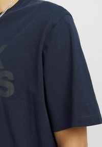 Tumesinine puuvillane T-särk, millel on lühikesed varrukad, ümar kaelus ja suur, kulunud trükk esiküljel. Sile tekstuur, mugav lõige.