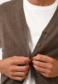 Brun cardigan i mjukt material med v-ringning, träknappar och en slät yta, lagd över en vit t-shirt.