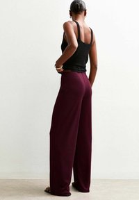 Une personne aux cheveux courts porte un débardeur noir et un pantalon large bordeaux, de dos contre un mur uni.