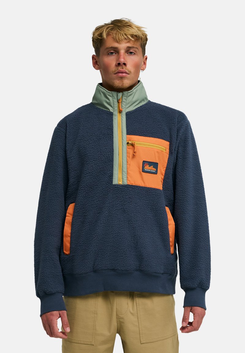 Φλις μπλούζα σε ναυτικό μπλε με ανοιχτό πράσινο γιακά και πορτοκαλί τσέπη στο στήθος. Διαθέτει φερμουάρ μέχρι τη μέση και υφή με ανάγλυφη επιφάνεια.