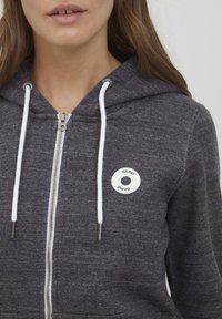 Mujer que lleva una sudadera con cremallera de color gris oscuro, con cordones blancos y un logo circular blanco de Oxmo en el lado izquierdo del pecho.