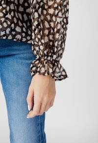 Blusa trasparente a stampa animale marrone con polsini arricciati; abbinata a jeans blu. Mano visibile con smalto chiaro appoggiata sulla coscia.