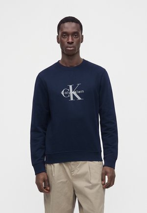 Mand iført en navyblå Calvin Klein sweatshirt og beige bukser, stående mod en ensfarvet lys baggrund, vendt fremad.
