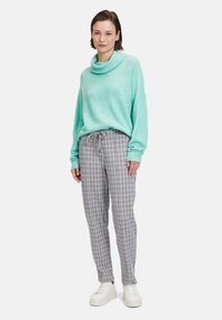 Licht turquoise cowl-neck sweater gecombineerd met geruite, broek met trekkoord in grijs en wit. Model draagt witte sneakers.