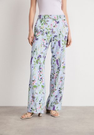Kvinde iført lyseblå bukser med vide ben og blomsterprint samt hvide sandaler med hæl og remme, stående på en grå gulv.