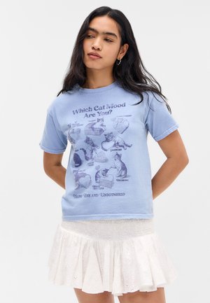 Vrouw draagt een lichtblauw t-shirt met vintage kattenillustraties en tekst, gecombineerd met een witte minirok met oogjes en ruches.