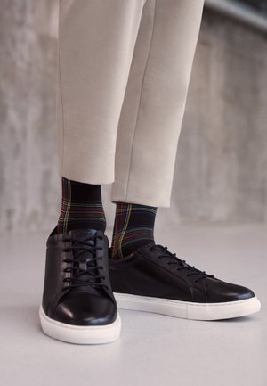Black leather sneakers with white rubber soles, featuring laces and a smooth finish. Paired with colorful plaid socks and beige trousers.