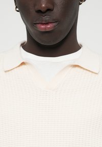 Lys beige strikket polo med v-hals, der har et tekstureret mønster og en blød finish, lagd over en hvid rundhalset skjorte.