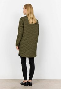 Lange olijfgroene gewatteerde jas met een ruitpatroon, lange mouwen en zijsplitsingen, gecombineerd met zwarte strakke broek en loafers.