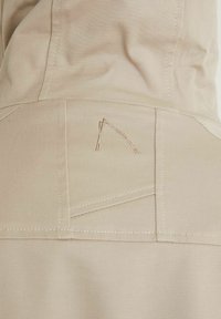 Närbild av beige tyg med detaljerad sömnad och en liten, abstrakt broderad logotyp i matchande trådfärg.