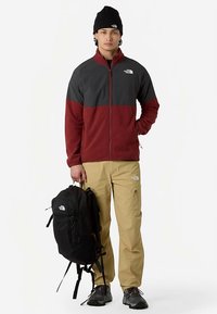 Czerwona i szara męska kurtka na zamek, zestawiona z beżowymi spodniami. Czarny plecak spoczywa na jednym ramieniu. Buty są czarne z fakturowaną podeszwą.