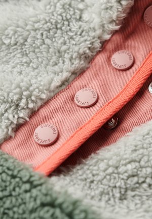 Detalle de chaqueta de forro polar con textura suave en gris, forro rosa y acentos en coral. Presenta botones redondos etiquetados como "Superdry" y un cierre a presión plateado.