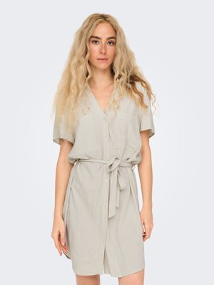 Femme aux longs cheveux blonds bouclés, portant une robe beige clair à manches courtes avec boutons et ceinture à la taille, debout devant un fond uni.