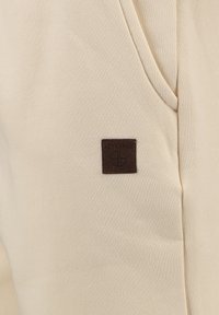 Beiger Stoff mit einer glatten Textur, versehen mit einem kleinen braunen Lederetikett, das mit "KEY LARGO" und einem Logo geprägt ist, auf einem schlichten Hintergrund.
