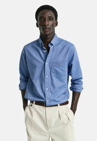 Lys blå skjorte med knapper laget av chambray-stoff, med en brystlomme og rullede ermer. Kombinert med kremfargede plisserte bukser.