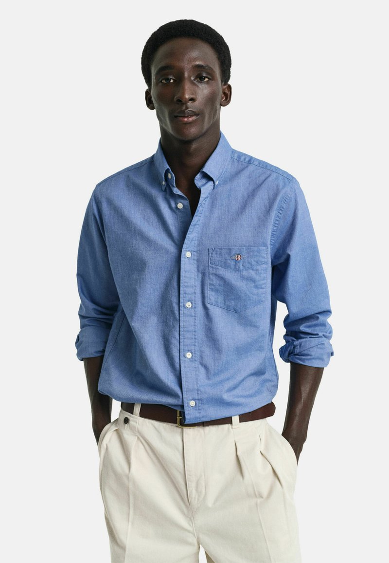 Lys blå skjorte med knapper laget av chambray-stoff, med en brystlomme og rullede ermer. Kombinert med kremfargede plisserte bukser.