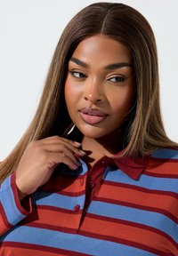 Czerwono-niebieska w paski koszulka polo z kołnierzykiem, z guzikami i miękką teksturą. Ręka modela spoczywa w pobliżu dekoltu.