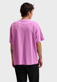 Osoba z krótkimi ciemnymi włosami, ubrana w prosty, luźny różowy T-shirt i czarne spodnie, zwrócona tyłem, z widocznymi tatuażami na obu ramionach.