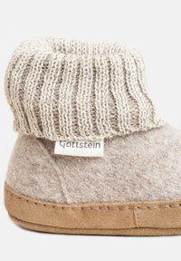Pantofole in lana beige con risvolto a coste e suola in suede, dotate di un piccolo etichetta in tessuto "Gottstein" sul lato.