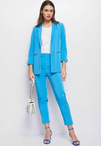 Blauwe blazer en bijpassende broek van lichte stof, gecombineerd met een wit T-shirt en blauwe haksandalen. Inclusief een witte handtas.