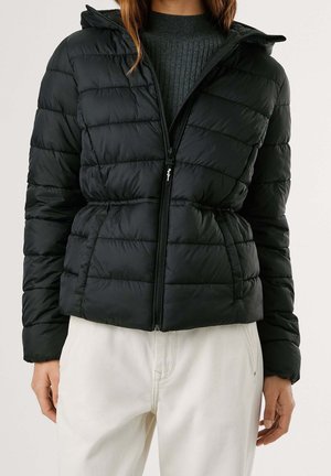Femme portant une veste matelassée noire à capuche par-dessus un pull côtelé gris foncé et un pantalon blanc, debout les mains détendues le long du corps.
