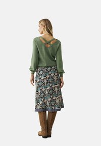 Jersey verde de punto con diseño en la parte trasera entrelazado, combinado con una falda estampada que presenta motivos florales. Botas marrones hasta la rodilla completan el look.