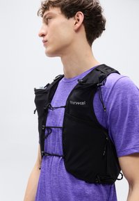 Μαύρο αμάνικο τζάκετ τρεξίματος με ρυθμιζόμενους ιμάντες και πολλαπλές τσέπες, φορεμένο πάνω από μοβ πουκάμισο. Διαθέτει ελαφρύ, ευέλικτο ύφασμα.