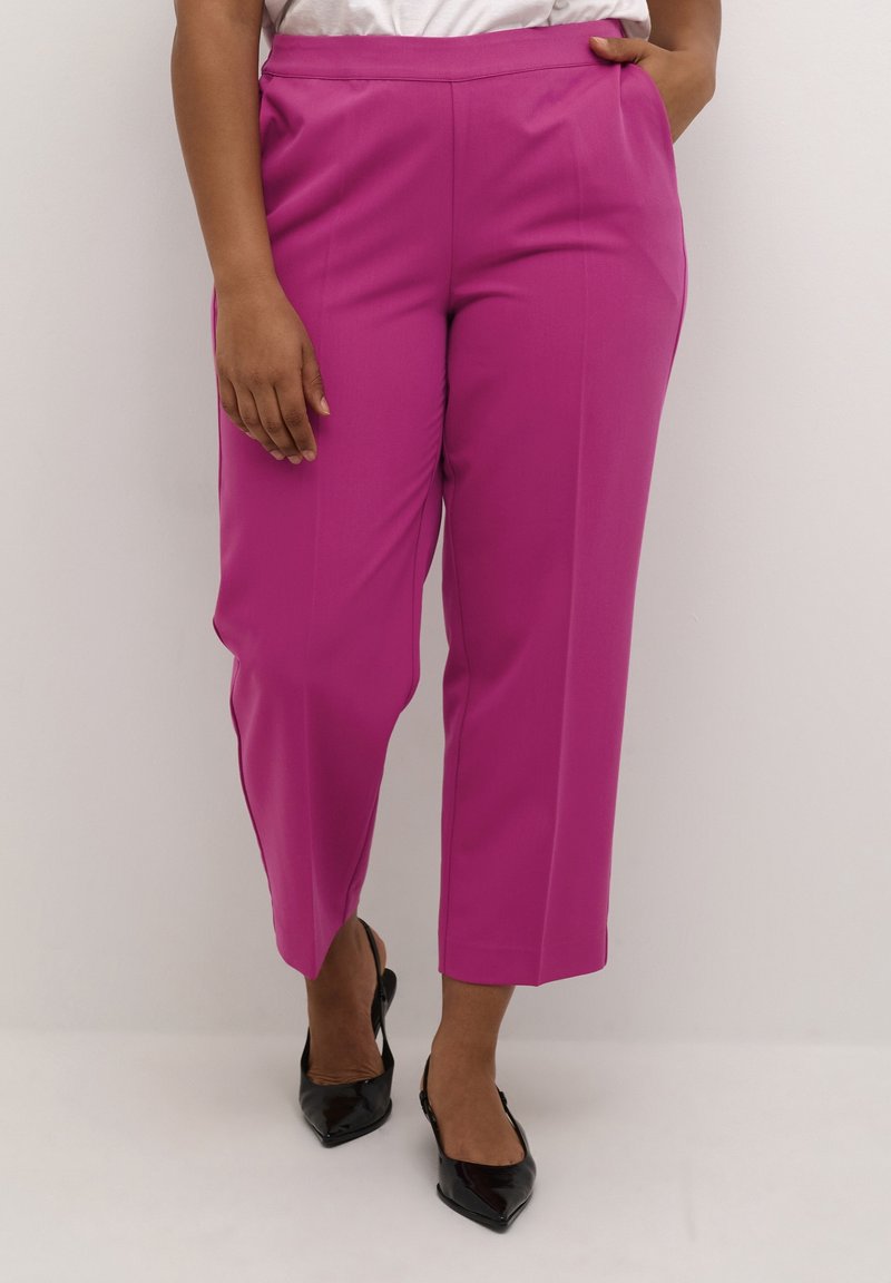 Femme portant un pantalon rose vif sur mesure longueur cheville avec poches latérales et des talons slingback noirs à bout pointu.