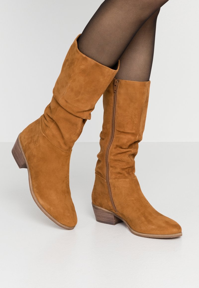Bottes montantes en daim marron avec un design souple, une fermeture éclair latérale et un talon bas en bois. La texture est douce et lisse.