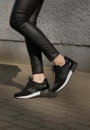 Persona que lleva pantalones de cuero negros y zapatillas negras con suelas blancas, de pie sobre una superficie exterior pavimentada cerca de una pared gris texturizada.