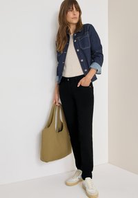 Giacca di jeans con polsini azzurri, maglione beige, pantaloni neri e una borsa verde. Indossa sneaker bianche con dettagli blu. Texture lisce.