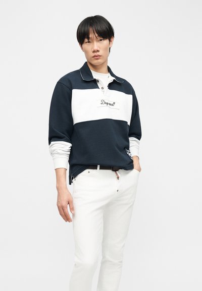 Jeune homme debout, une main dans la poche, portant un polo à manches longues bleu marine et blanc ainsi qu'un pantalon blanc, devant un fond uni.