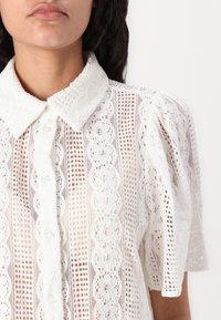 Blusa bianca accorciata con una sovrapposizione in pizzo testurizzato, chiusura con bottone e colletto classico, con maniche corte a sbuffo e delicati motivi.