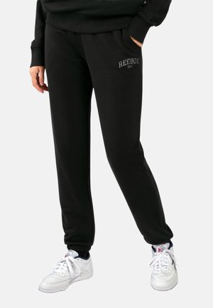 Persoon die zwarte Reebok-trainingsbroek met zakken en witte Reebok-sneakers draagt tegen een effen lichte achtergrond.