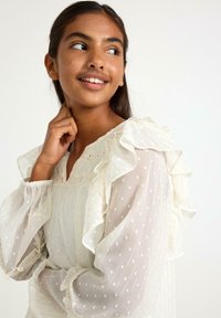 Ung kvinde med mørkt hår iført en hvid, gennemsigtig langærmet bluse med flæser og prikker, smilende og kiggende til siden.